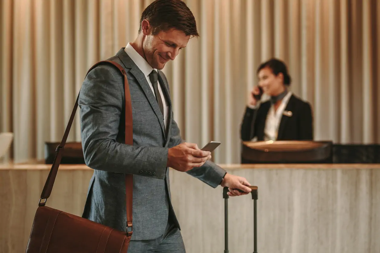 Hotel biznesowy i biznesmen spacerujący po holu z walizką i korzystający ze smartfona. Mężczyzna podróżujący służbowo w holu hotelu z telefonem komórkowym i bagażem.